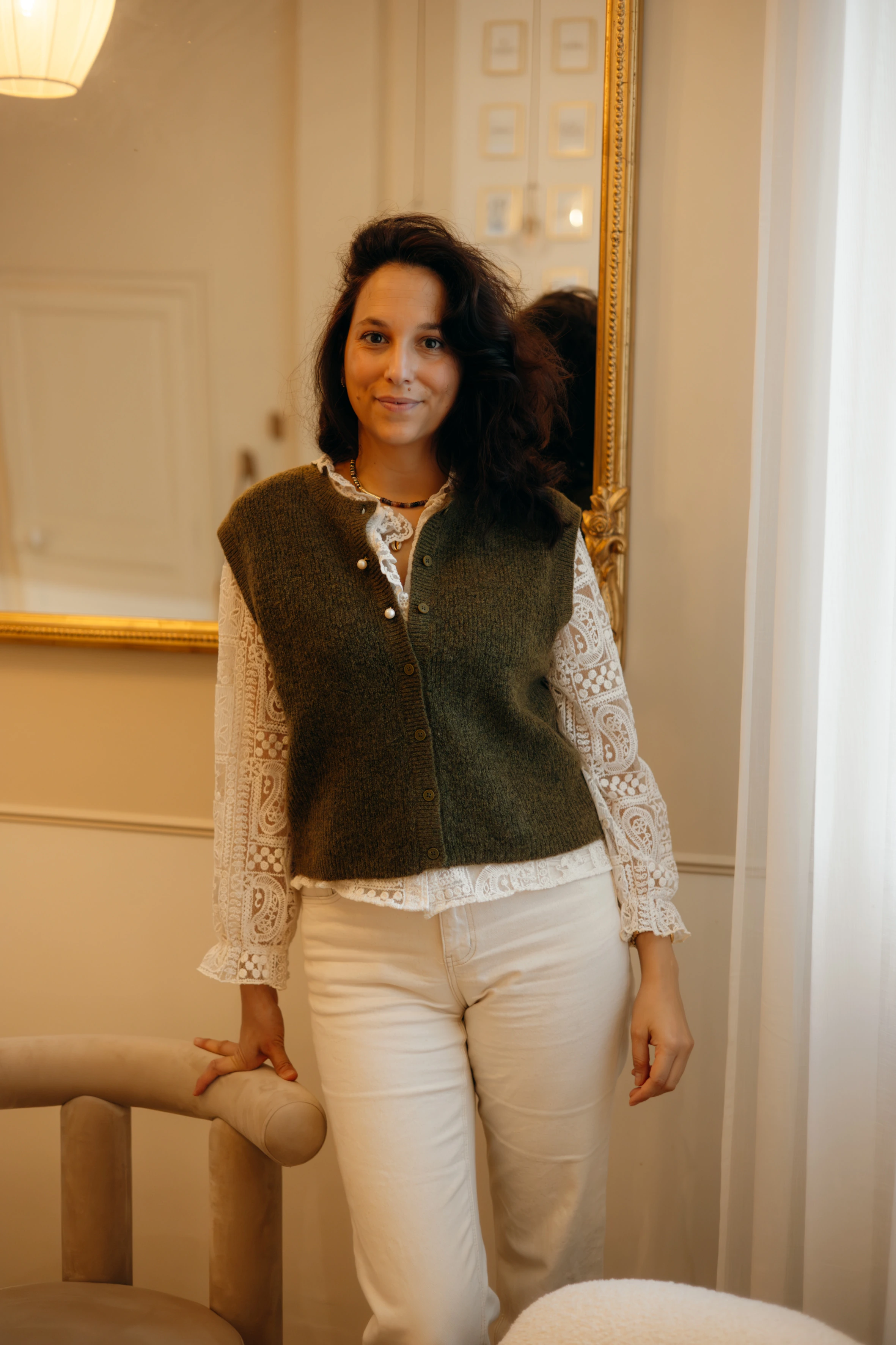 Julie Clément, debout près d’un miroir, en gilet vert et chemisier en dentelle.
